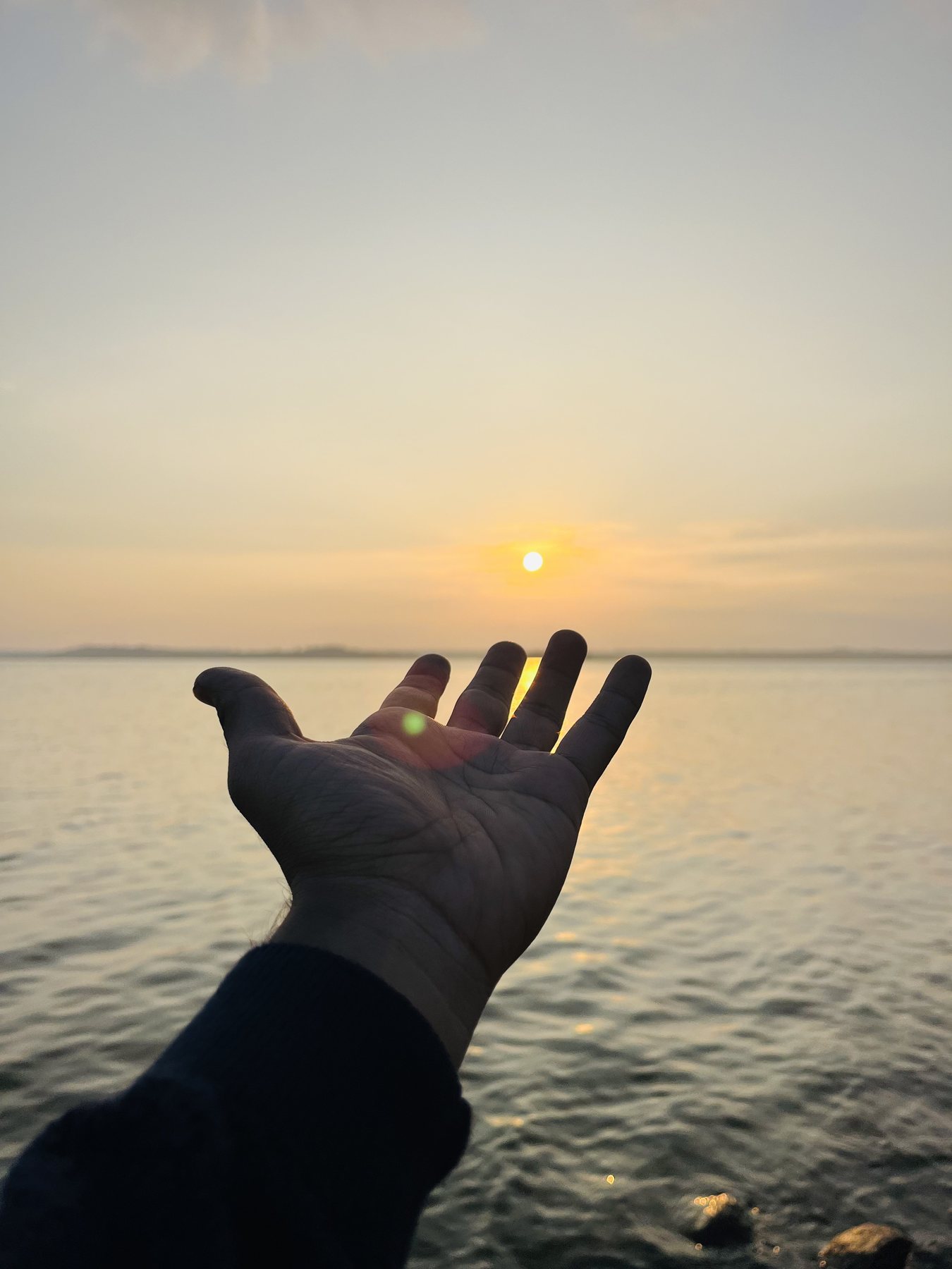A hand reaching toward the setting sun above the sea